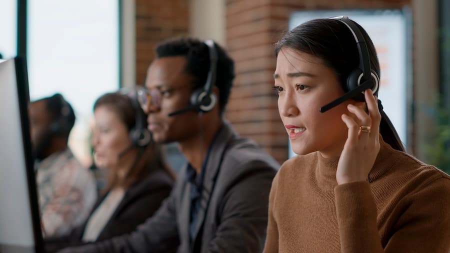Three customer service representatives wearing headsets work at computers in a modern office. The scene is focused, professional, and collaborative.