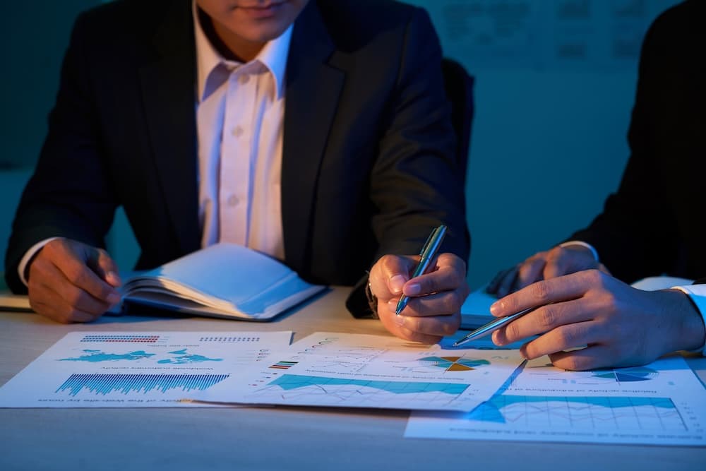 Two individuals in suits analyze graphs and charts on a table under dim lighting, creating a focused and professional atmosphere.