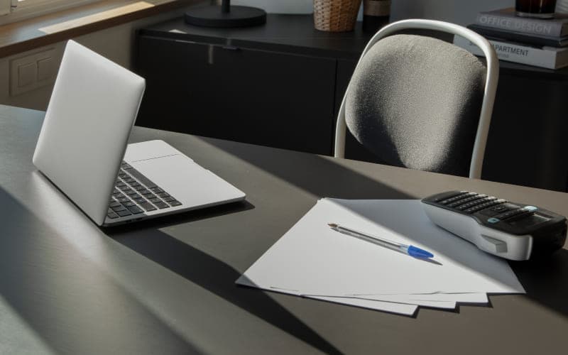 A tidy desk with a laptop, stacked papers, a blue pen, and a label maker. Sunlight casts long shadows, creating a calm and organized atmosphere.