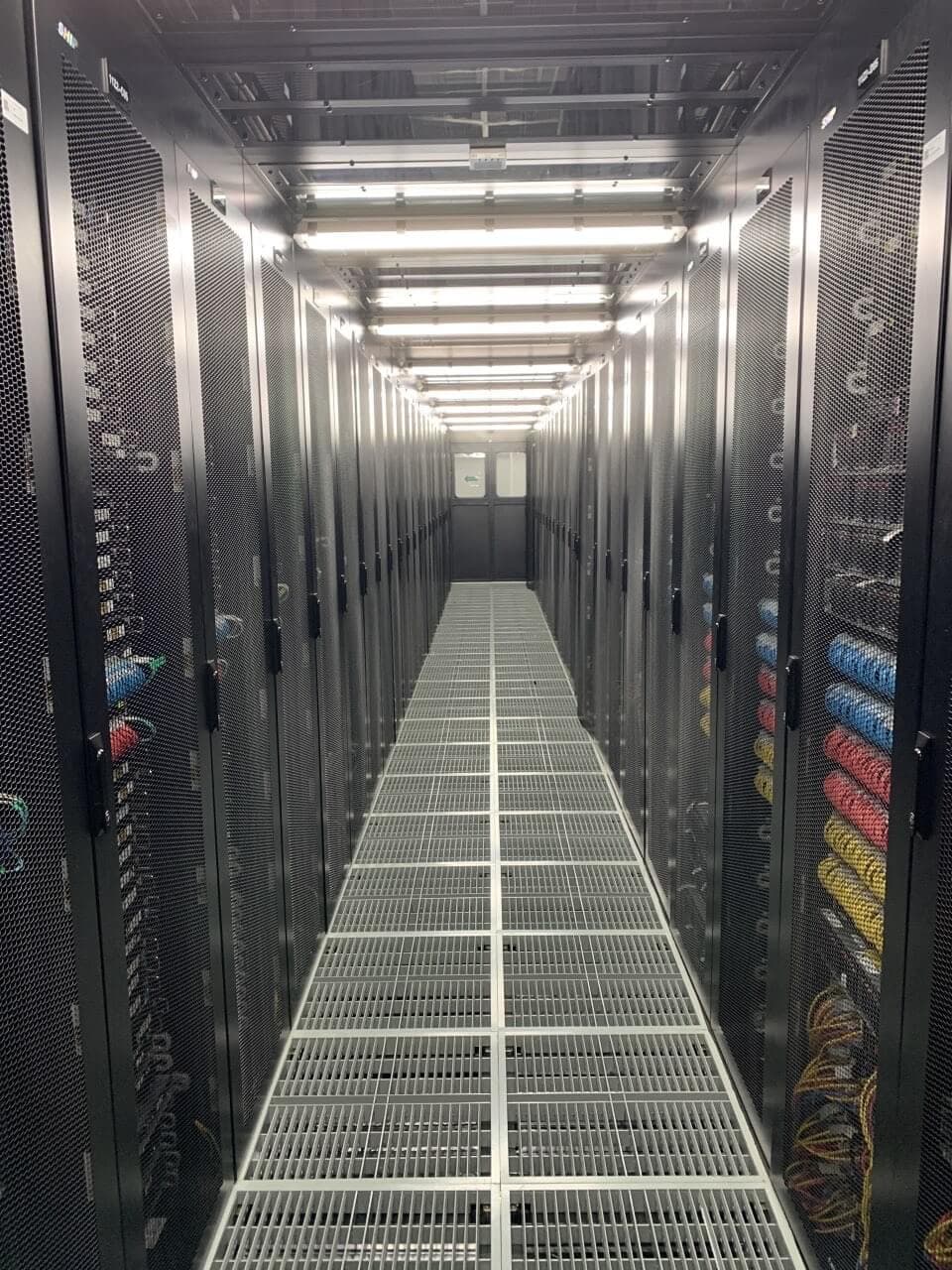 Long server corridor with raised floor grating, parallel server cabinets, and color-coded cable management systems visible through mesh doors