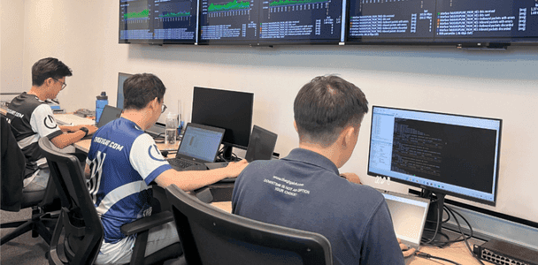 Three people in a network operations center on laptops and a desktop showing code. Multiple screens display data above them, suggesting a focused, collaborative atmosphere. Three people in a network operations center on laptops and a desktop showing code. Multiple screens display data above them, suggesting a focused, collaborative atmosphere.
