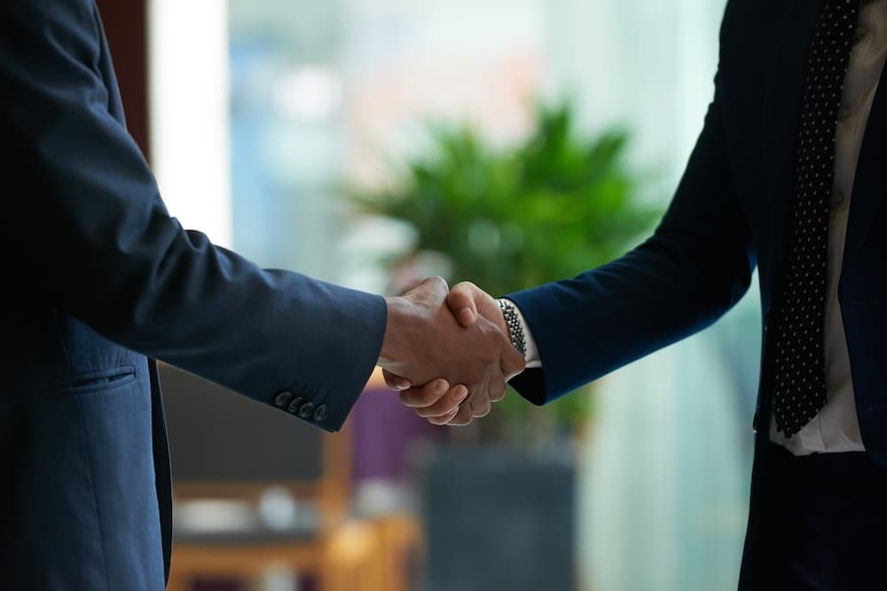 Two individuals in dark suits shake hands, symbolizing a formal agreement or partnership. Two individuals in dark suits shake hands, symbolizing a formal agreement or partnership.