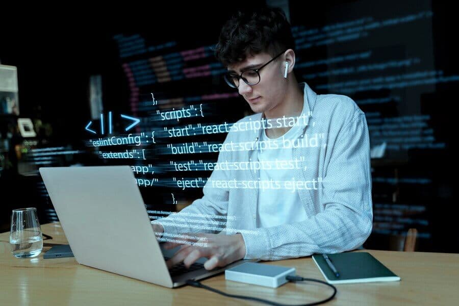 Young man wearing earphones and glasses works on a laptop with programming code overlayed. Young man wearing earphones and glasses works on a laptop with programming code overlayed.