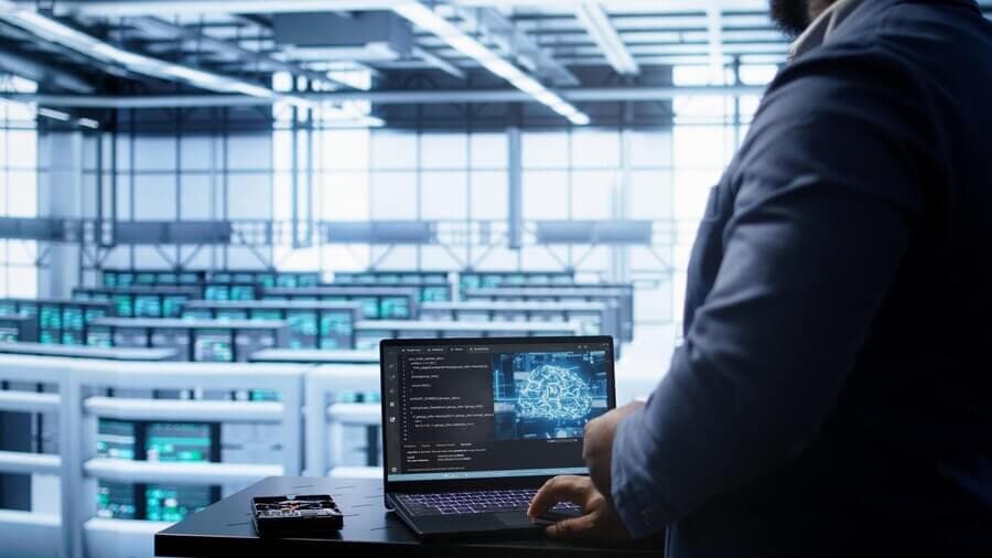 A person stands working on a laptop showing code and a digital brain diagram, set against a data center with rows of servers. A person stands working on a laptop showing code and a digital brain diagram, set against a data center with rows of servers.