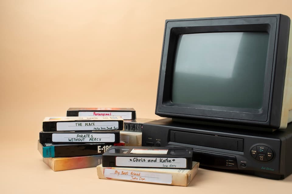 Vintage setup with a CRT TV and VHS player on the right. To the left, a stack of labeled VHS tapes. Vintage setup with a CRT TV and VHS player on the right. To the left, a stack of labeled VHS tapes.