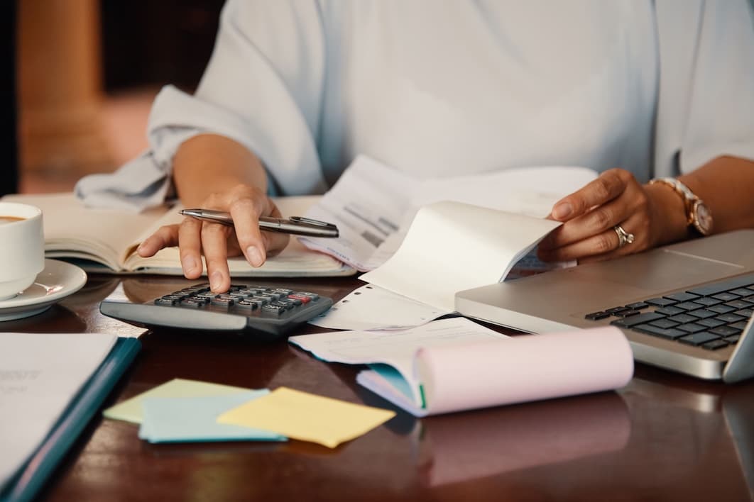 A person works at a desk with a calculator, pen, and laptop, surrounded by documents and a coffee cup, conveying focus and productivity in an office setting. A person works at a desk with a calculator, pen, and laptop, surrounded by documents and a coffee cup, conveying focus and productivity in an office setting.