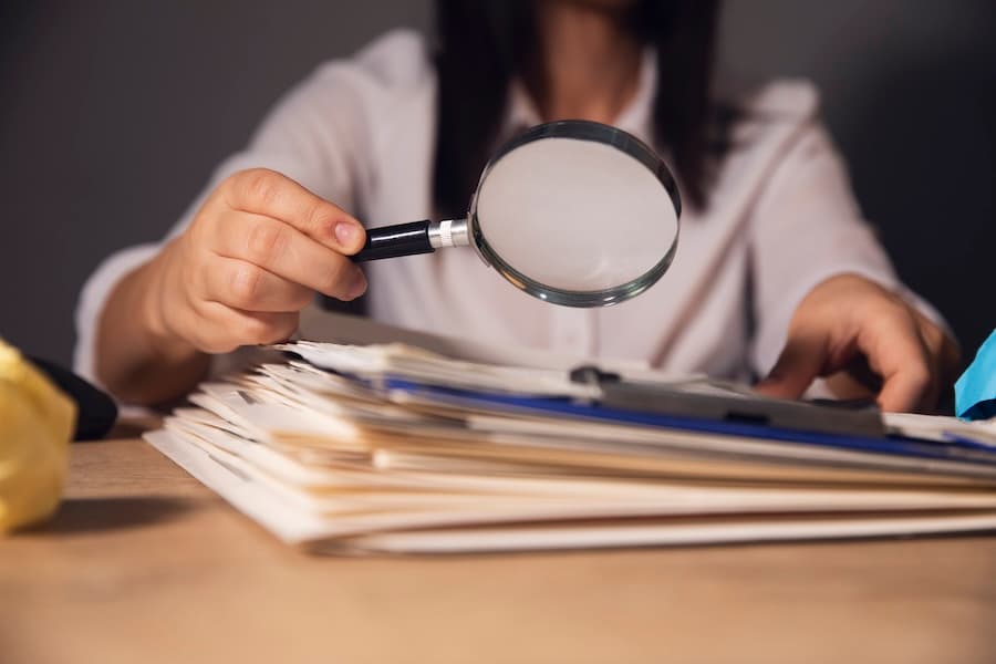 A person examining documents with a magnifying glass, representing detailed investigation or auditing. A person examining documents with a magnifying glass, representing detailed investigation or auditing.
