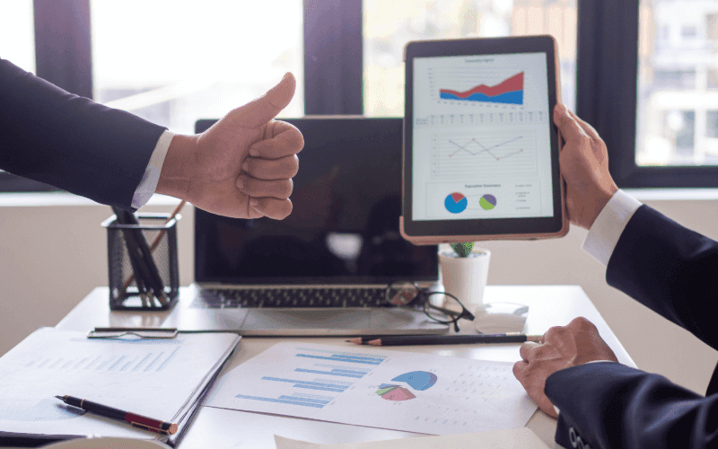 Two businesspeople in suits at a desk reviewing charts on a tablet. One shows a thumbs-up, suggesting positivity. Papers and a laptop are visible. Two businesspeople in suits at a desk reviewing charts on a tablet. One shows a thumbs-up, suggesting positivity. Papers and a laptop are visible.