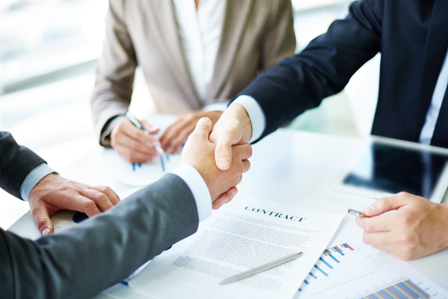 Two businesspeople in suits handshake over a contract on a table, with a pen and charts visible. Two businesspeople in suits handshake over a contract on a table, with a pen and charts visible.